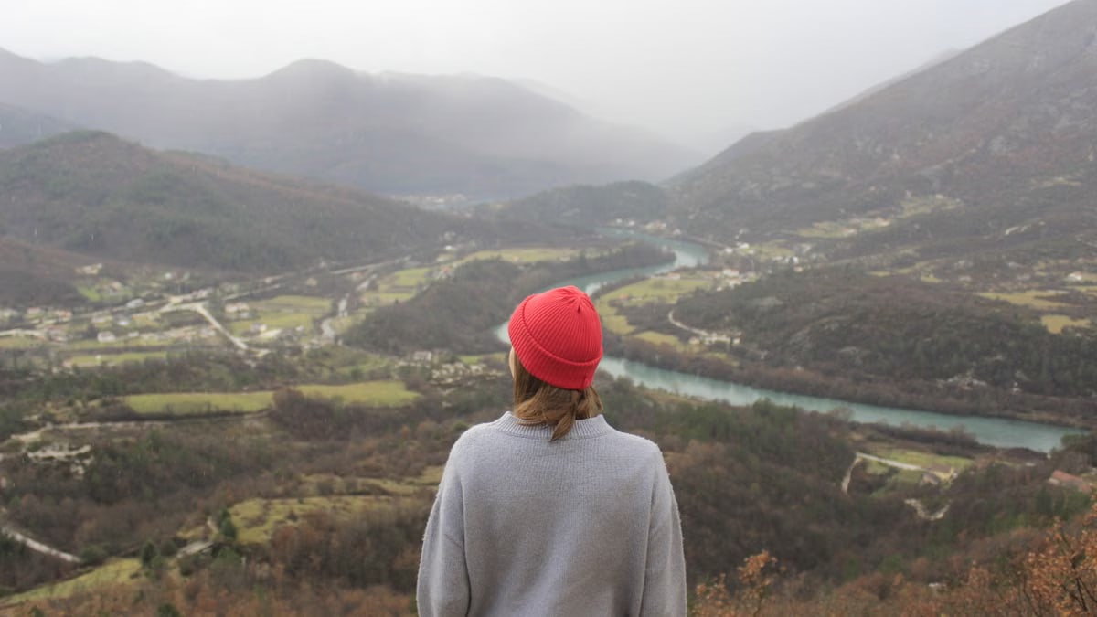 A woman in a red hat overlooking a vast mountain valley and winding river.